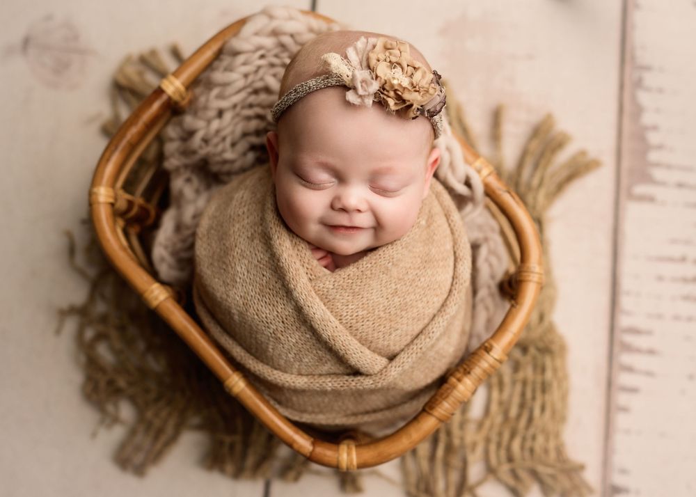 Smiling newborn baby wrapped in tan swaddle with gold flower headband resting in wicker basket.