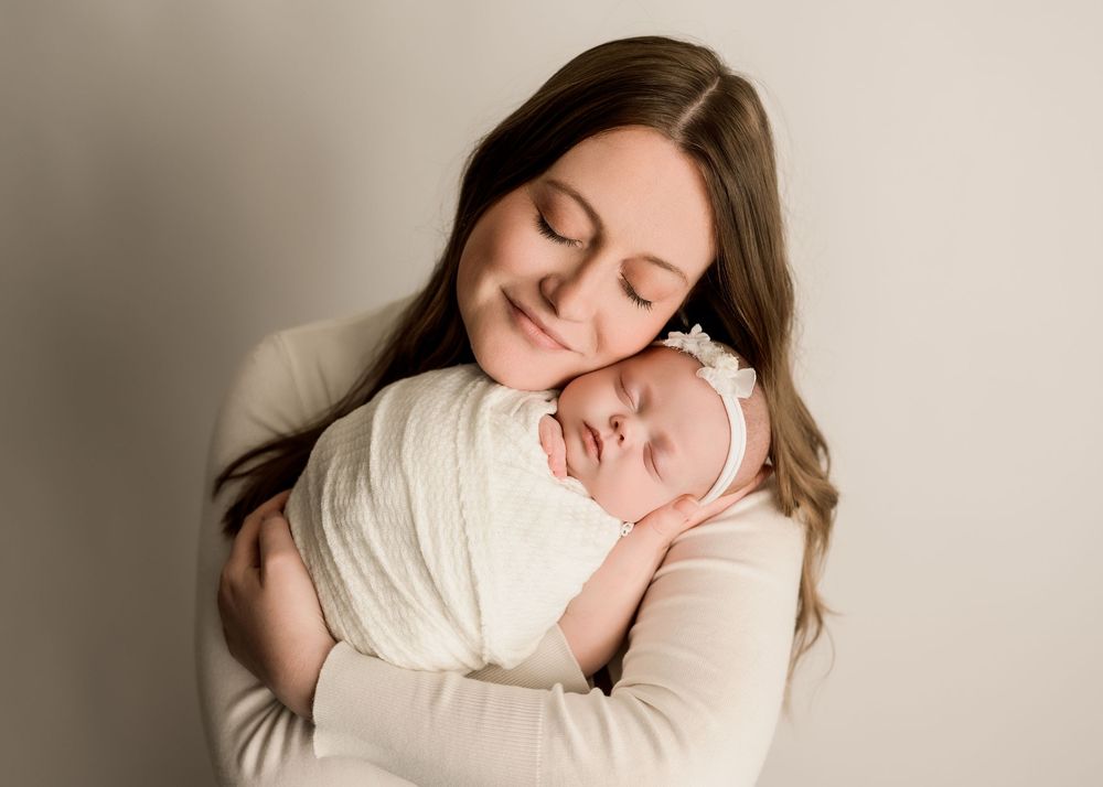 Mother with eyes closed tenderly holds swaddled newborn baby girl against her cheek in white outfit.
