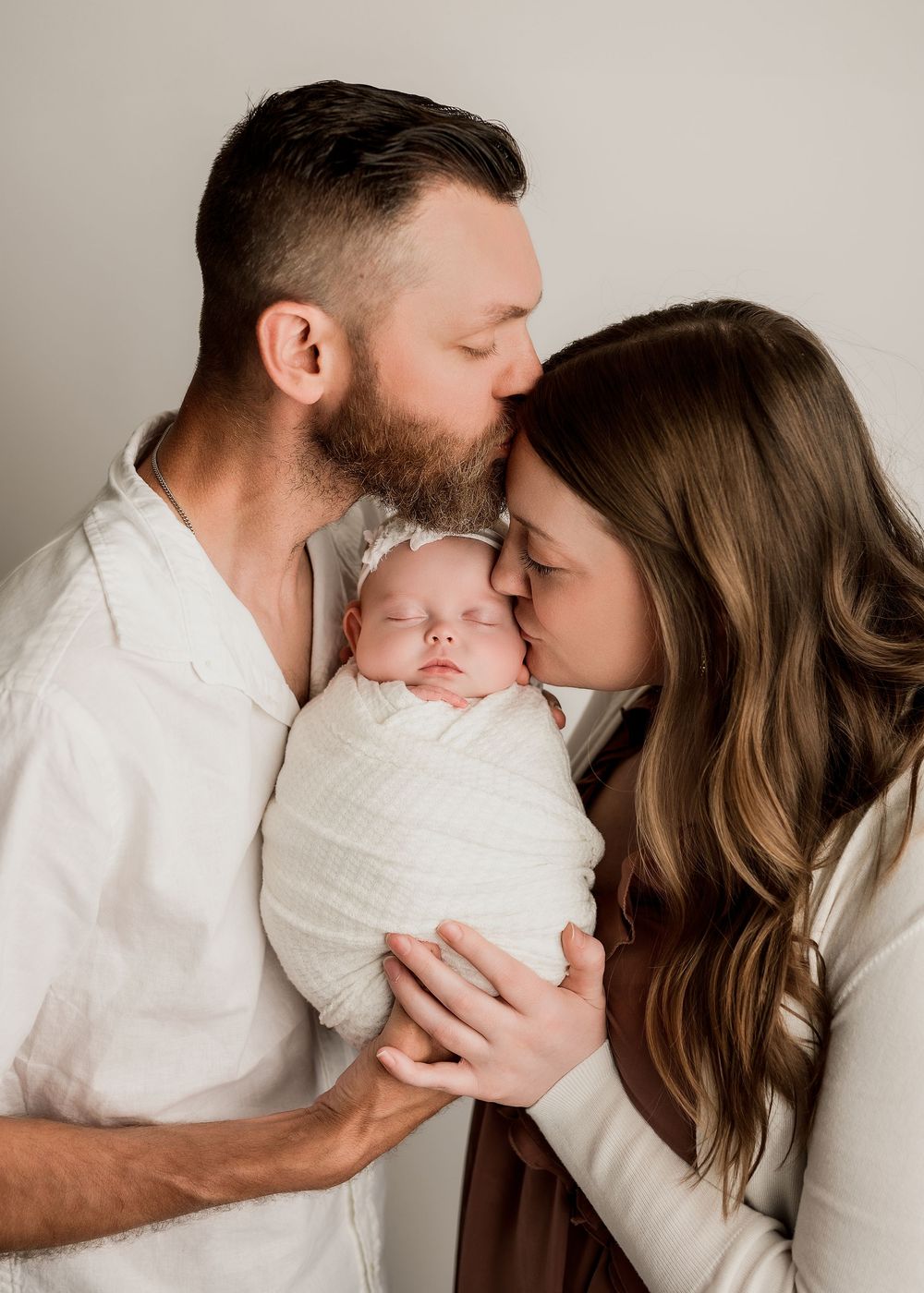 Parents lovingly kiss their swaddled newborn baby girl on the head against neutral background.