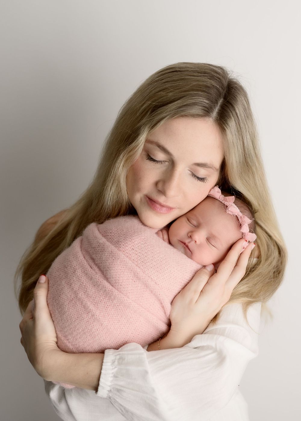 Mother tenderly holds sleeping pink-swaddled newborn baby girl close to her cheek in a soft studio portrait.