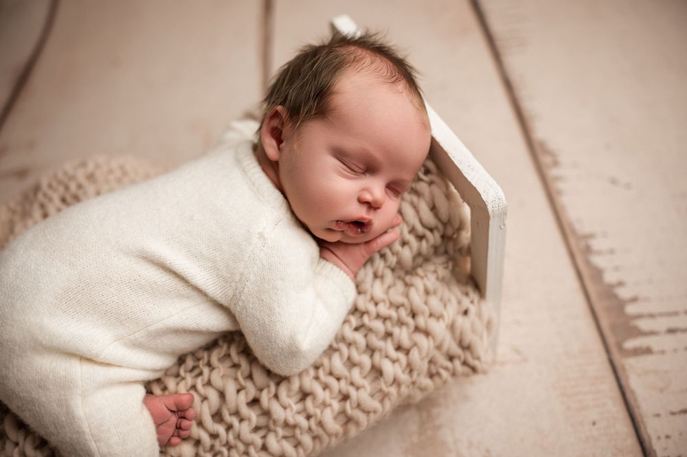 Sleeping newborn baby in cream knit outfit resting on chunky knit blanket on wood floor.
