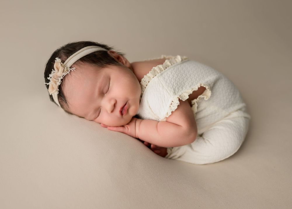 Sleeping newborn baby in white lace romper with pearl headband on neutral background.