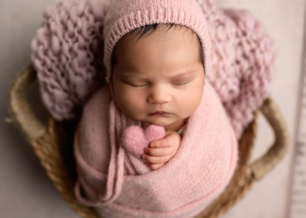 Newborn baby swaddled in pink knit wrap with matching bonnet holding pink heart in wicker basket.