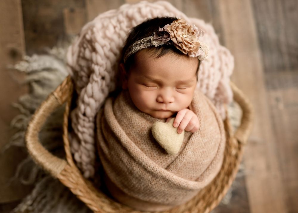Sleeping newborn wrapped in beige knit blanket inside wicker basket holding a small heart prop.