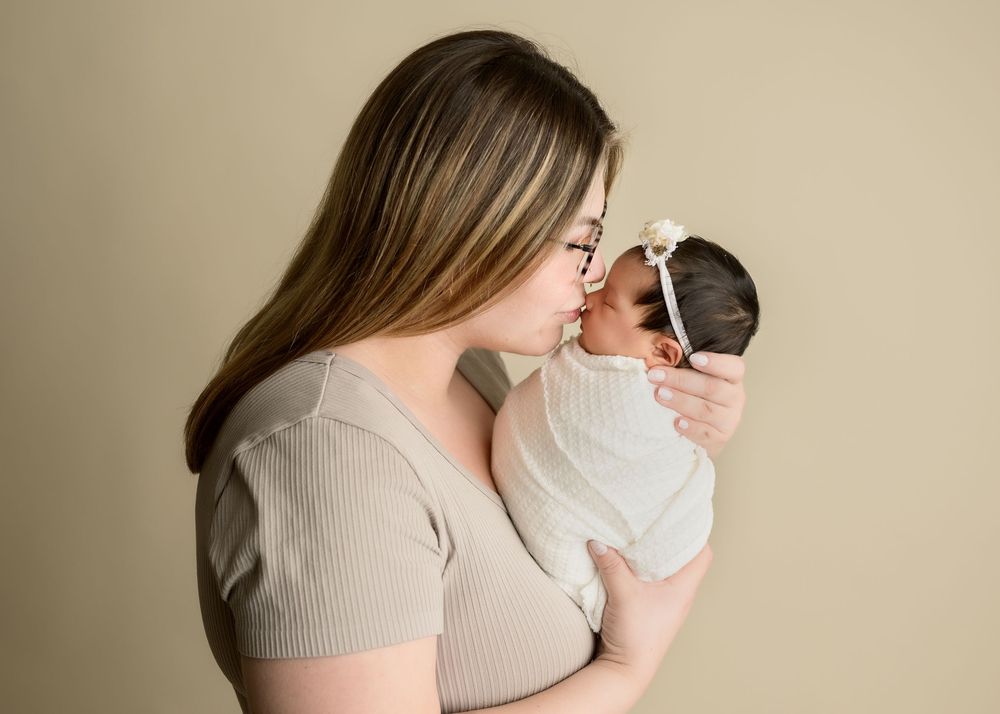 Mother gently kissing her swaddled newborn baby against a soft neutral beige studio background.