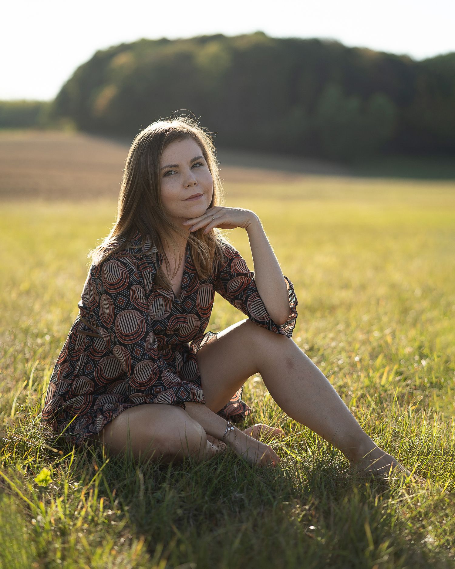 Young woman in patterned dress sitting in sunlit green field at golden hour.