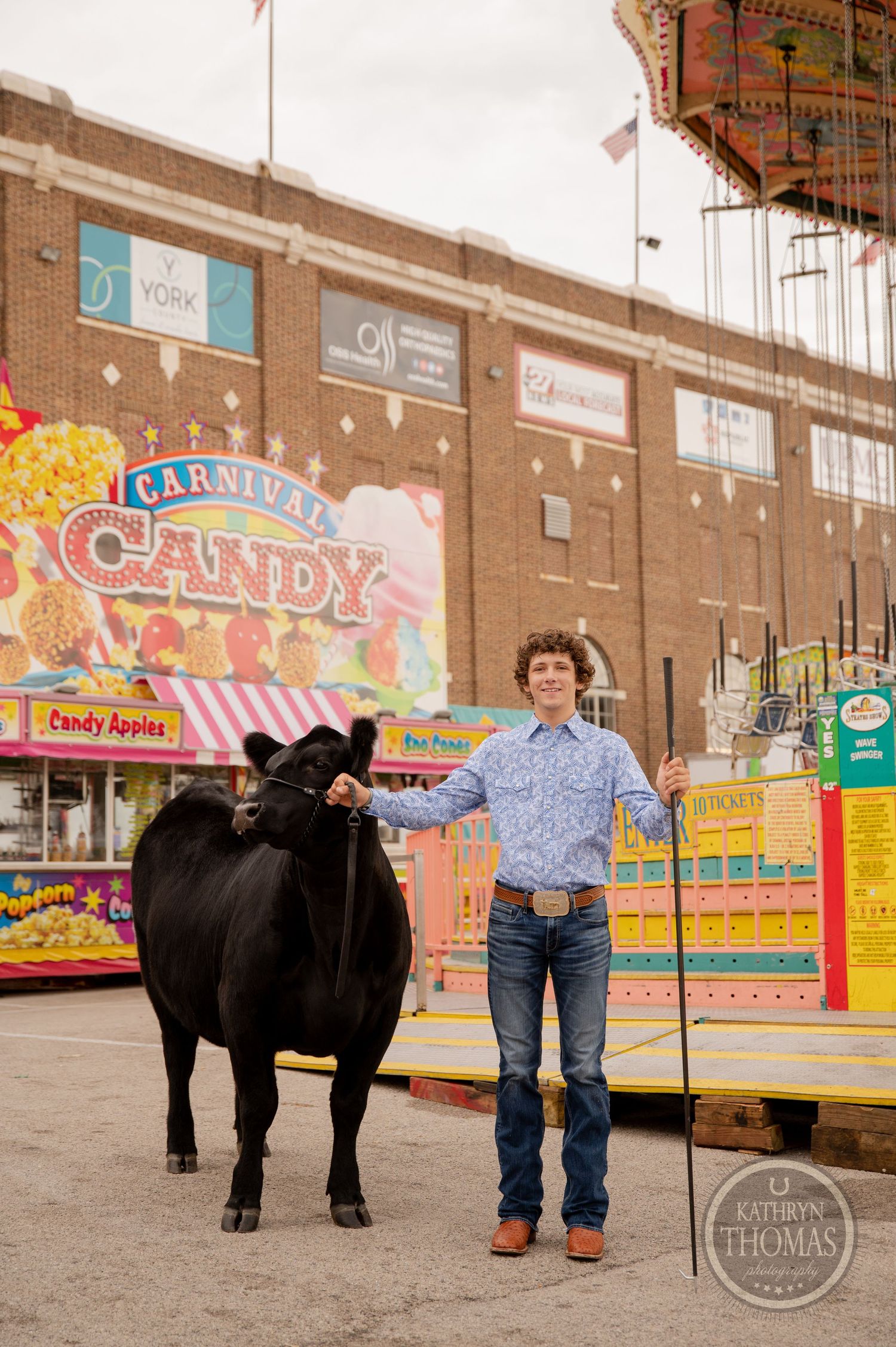 A person poses with a black show cow in front of carnival candy stands and game booths.