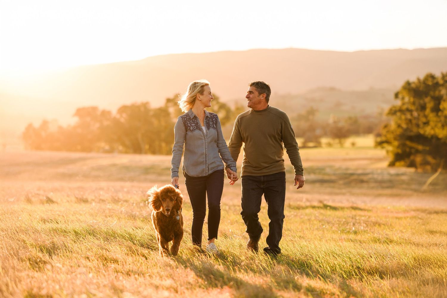 Couple poses with their golden retriever dog in a sunny outdoor field at golden hour.