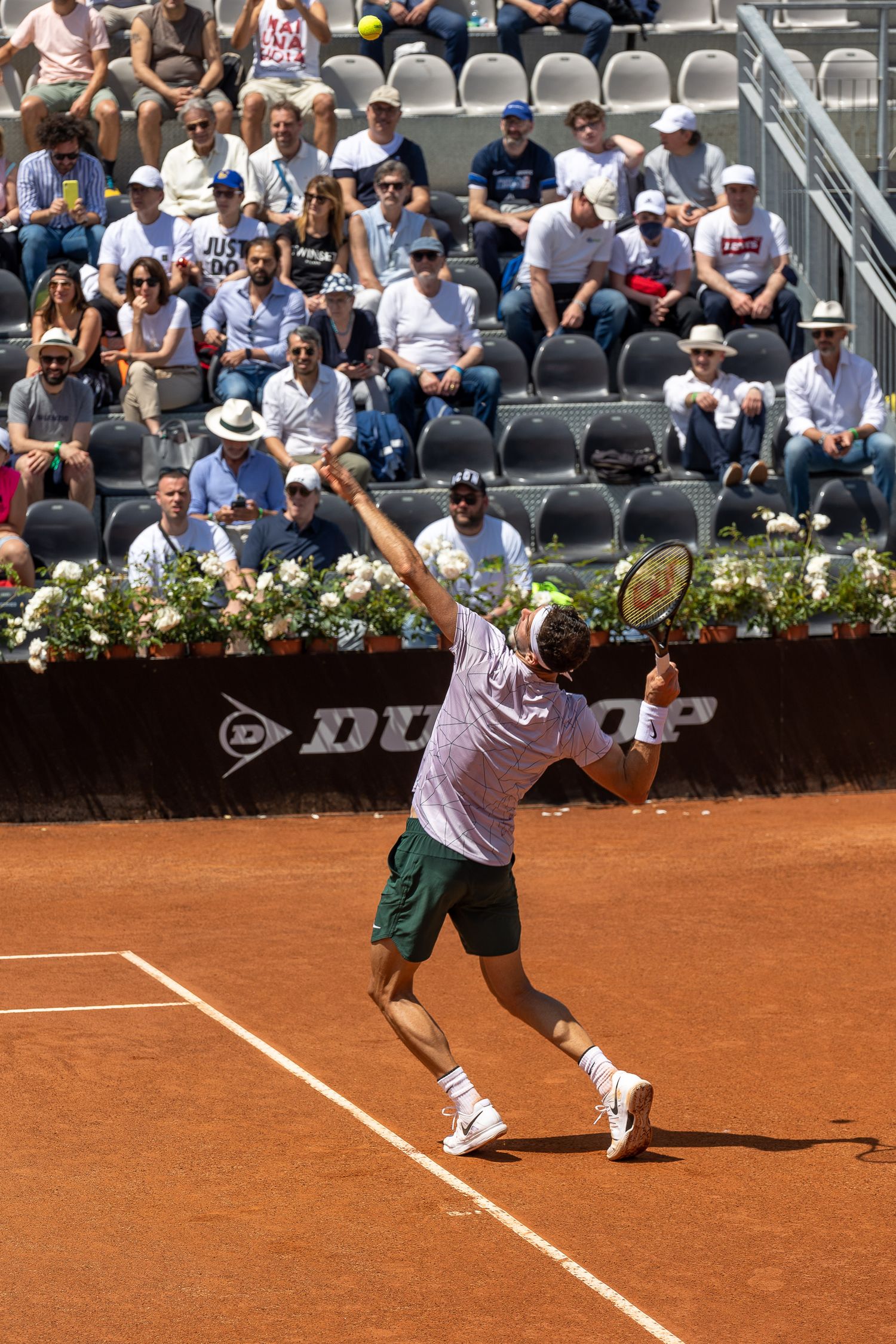 Tennis player serves ball on clay court during tournament match with spectators watching.