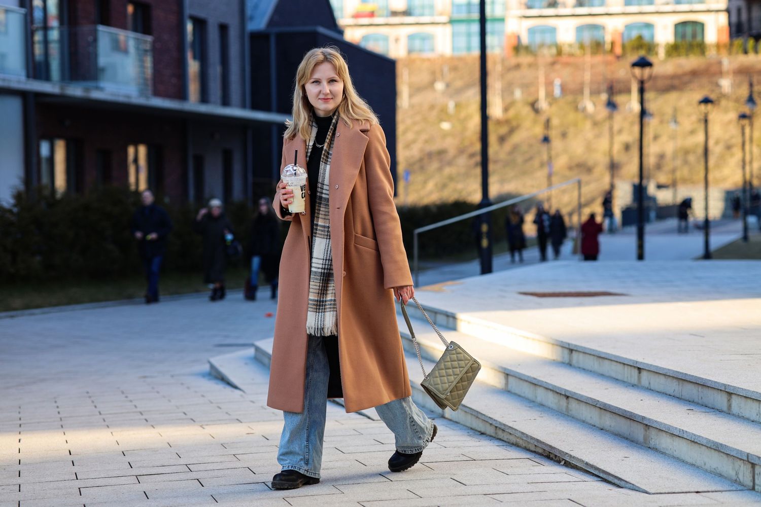 A fair-haired girl in a brown coat walks through Vilnius with a cup of coffee from VERO CAFE