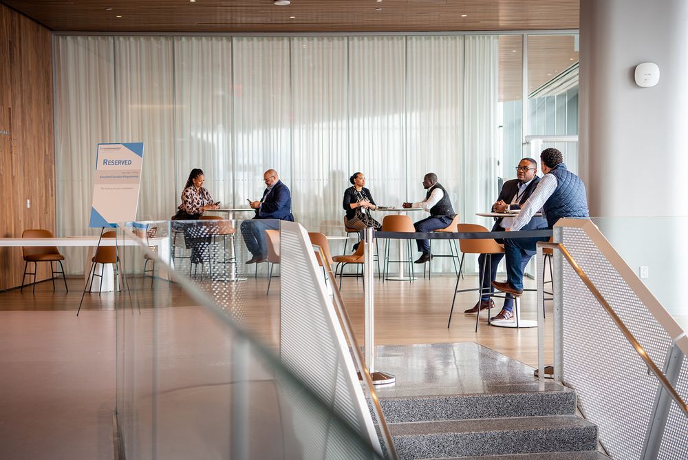 A group sits in a modern office lounge area with glass walls and contemporary furniture for a casual meeting.
