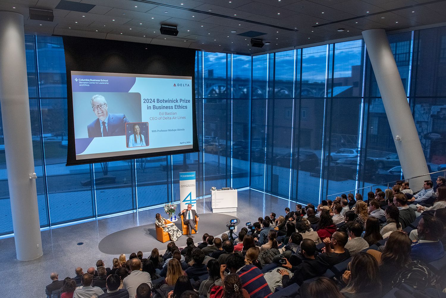 A large audience watches a presentation in a modern conference room with floor-to-ceiling windows and blue lighting.