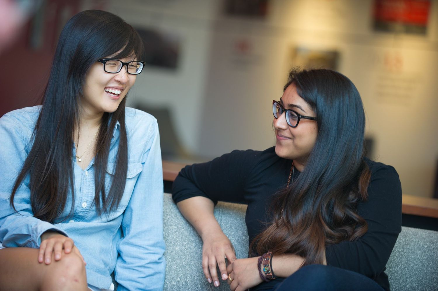 Two young women with long dark hair and glasses having an animated conversation while sitting together.