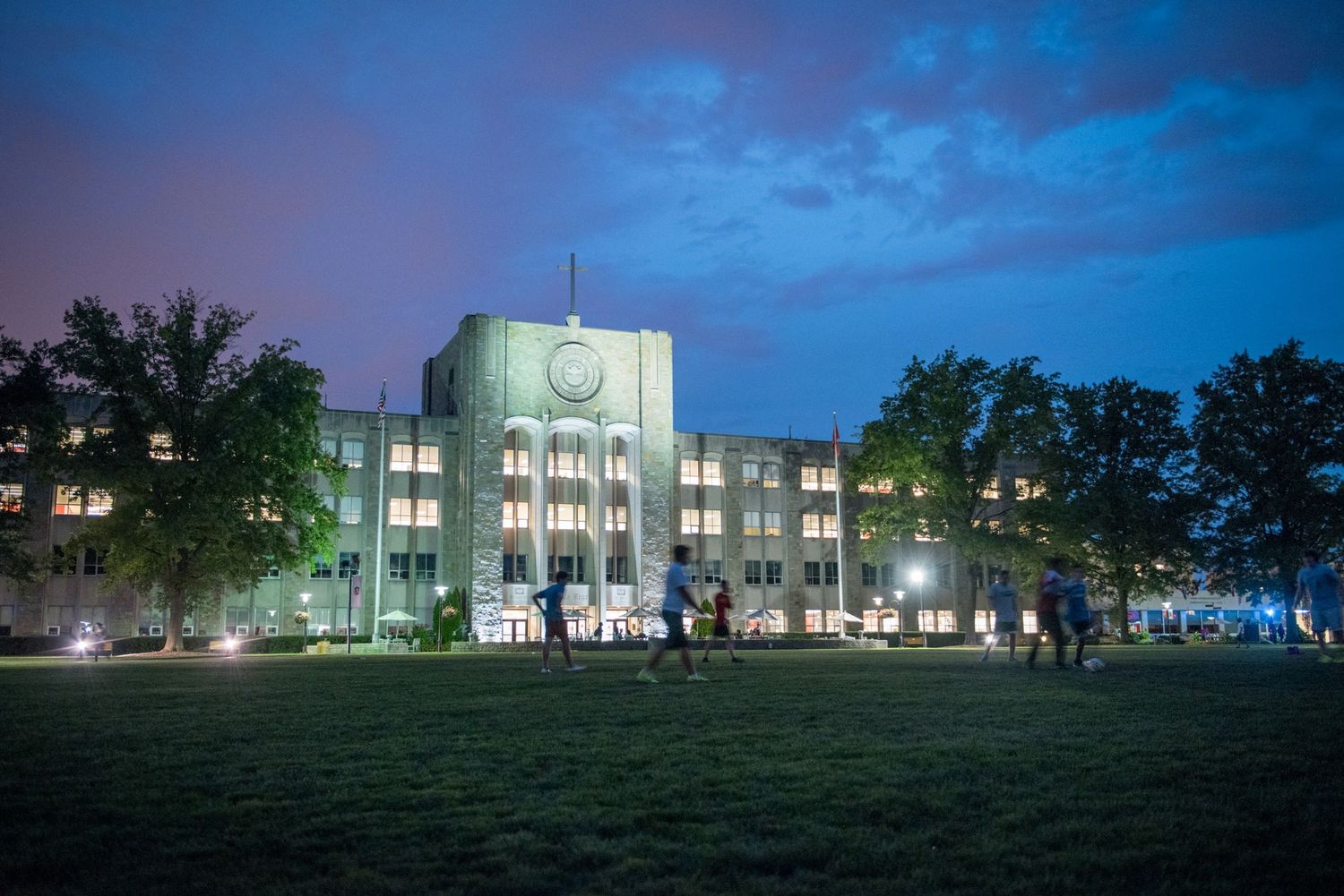 White illuminated academic building with glowing clock tower on a college campus at dusk with blue evening sky.