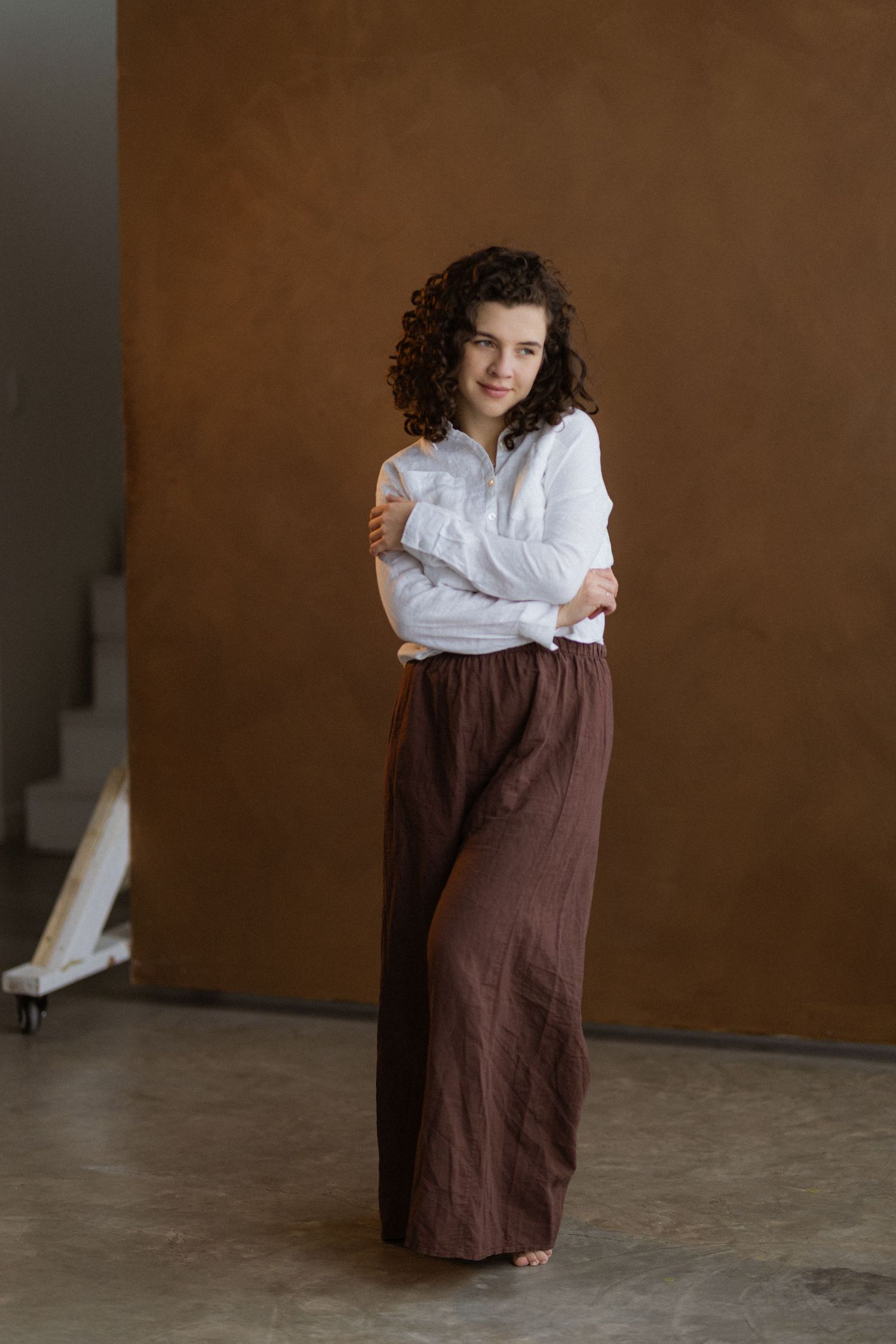 Stylish portrait of someone in a crisp white button-down shirt and flowing brown linen palazzo pants against a brown backdrop.