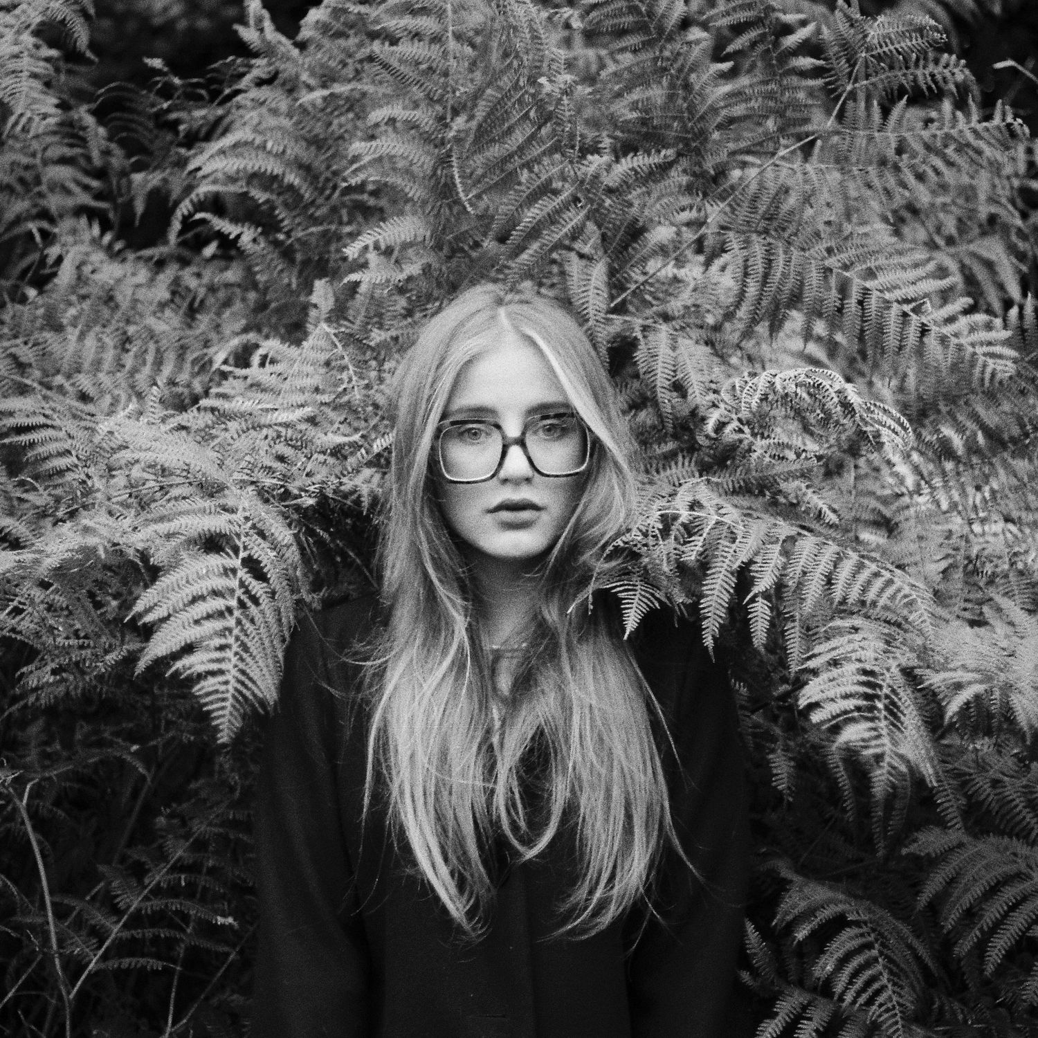 Portrait among ferns shows person in dark clothing with glasses against black and white nature backdrop.