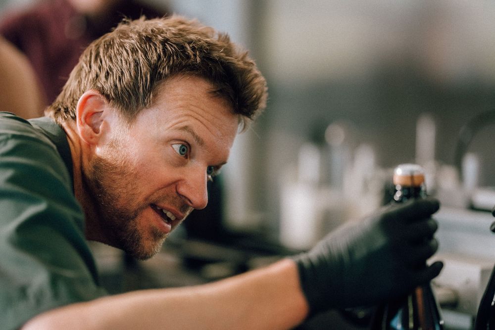 A person wearing a green jacket and black gloves smiles while working with equipment on a blurred background.