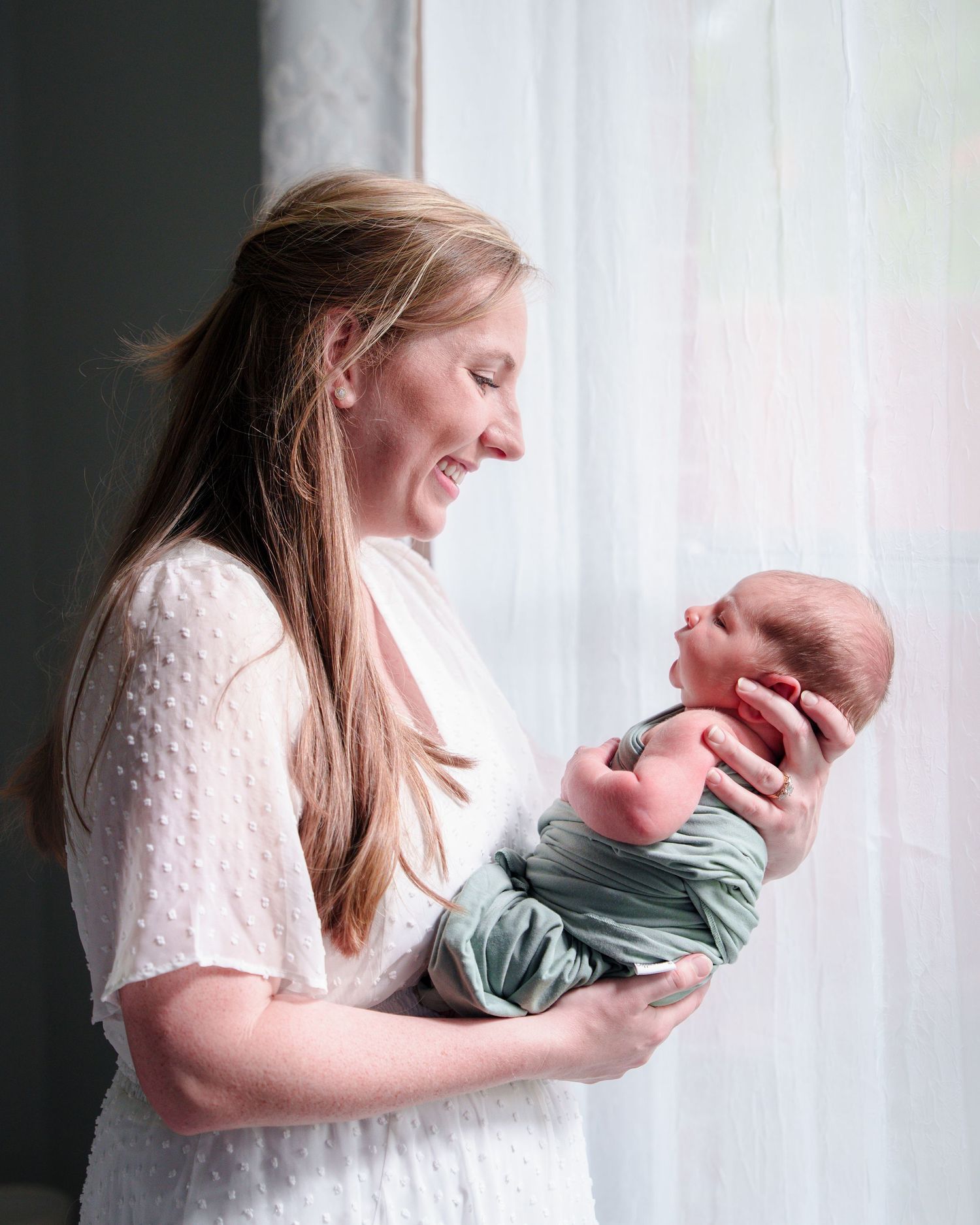 Newborn photo session at home natural light  Keller TX baby boy green  mom holding the baby by the window kissing