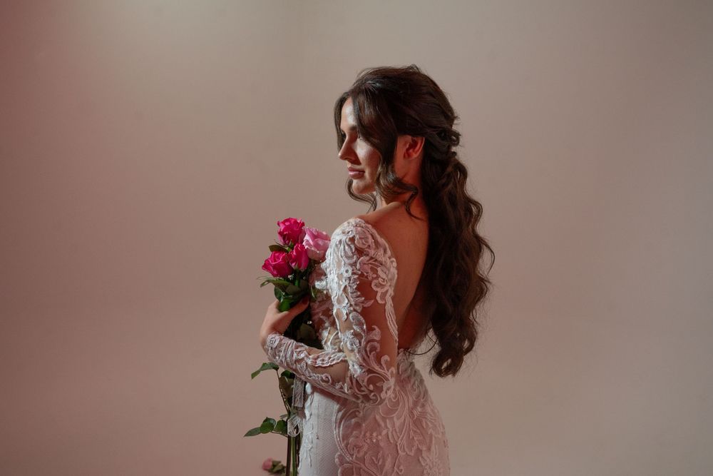 Bride in lace backless wedding gown holds pink roses in soft studio lighting.