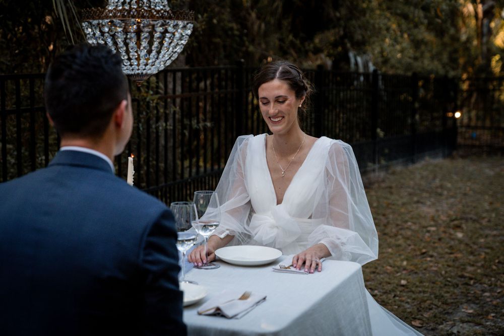 Smiling bride in sheer white gown sits across groom at outdoor wedding reception table.