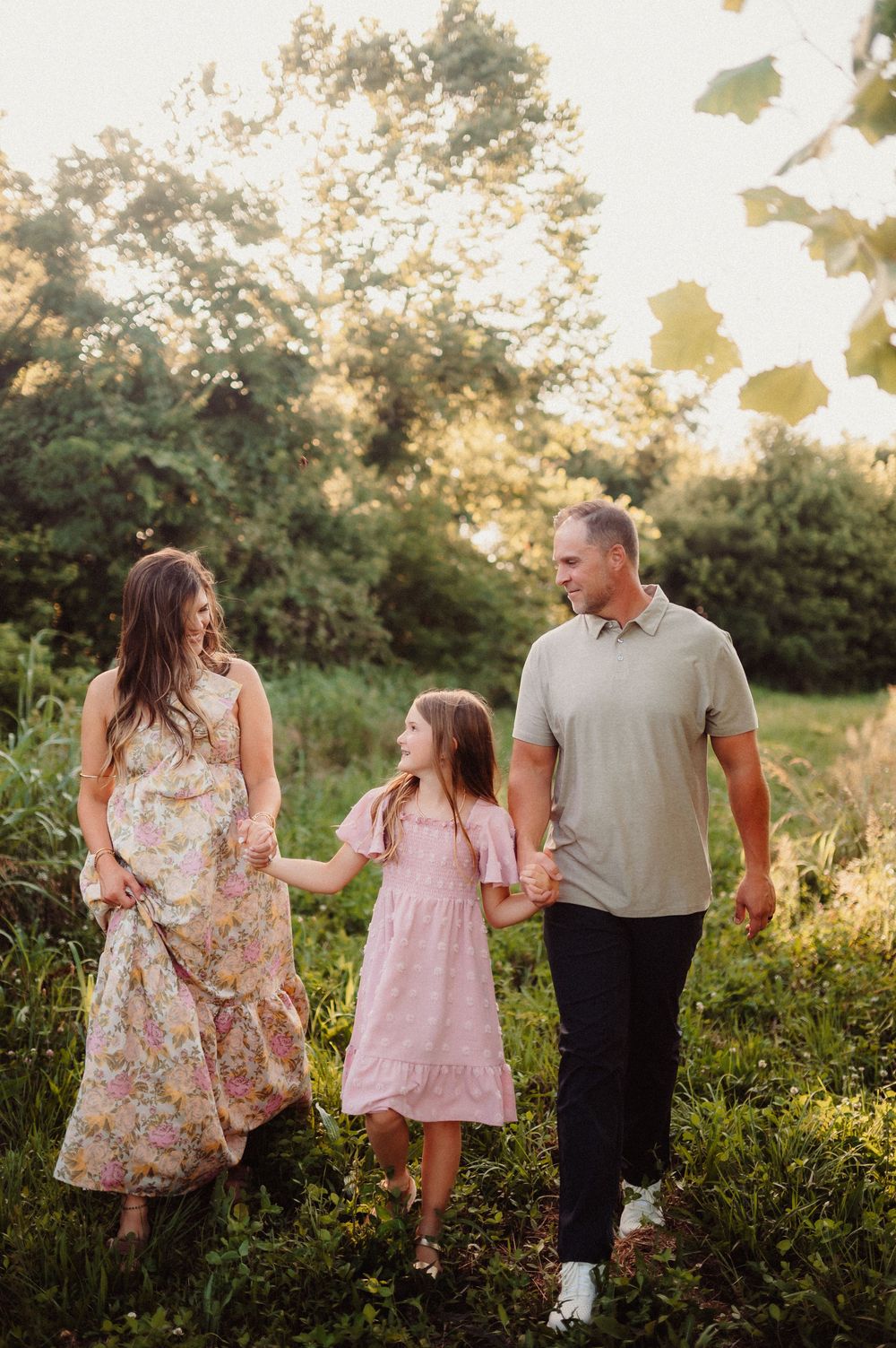 Family walks together in a scenic field at golden hour while wearing floral dress and casual attire.