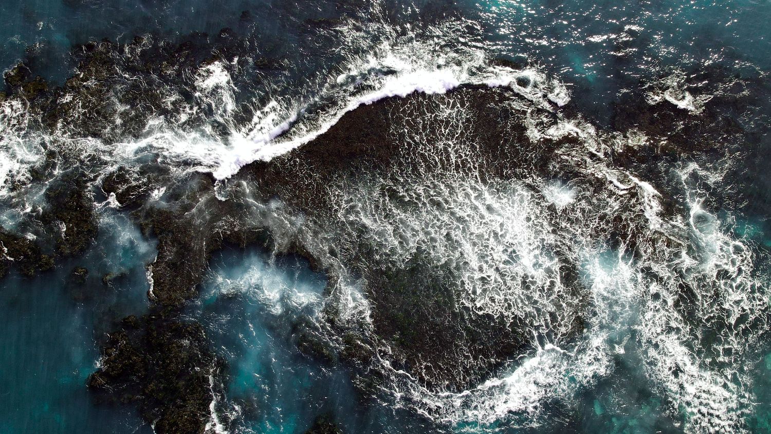 Aerial view of dramatic ocean waves crashing against dark rocky coastline.
