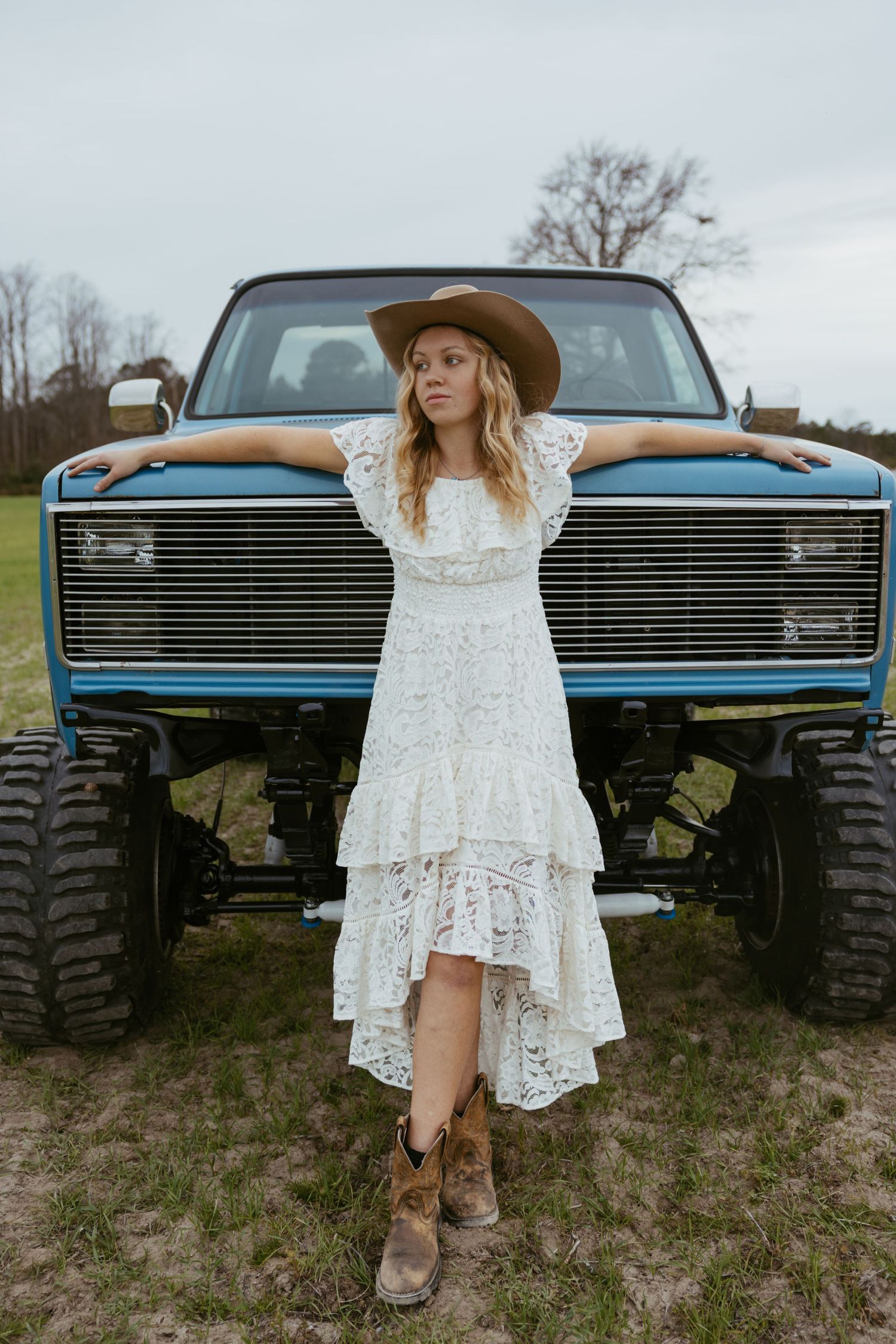A white lace dress and cowboy hat contrasts with a lifted blue vintage truck with large off-road tires in a rural setting.