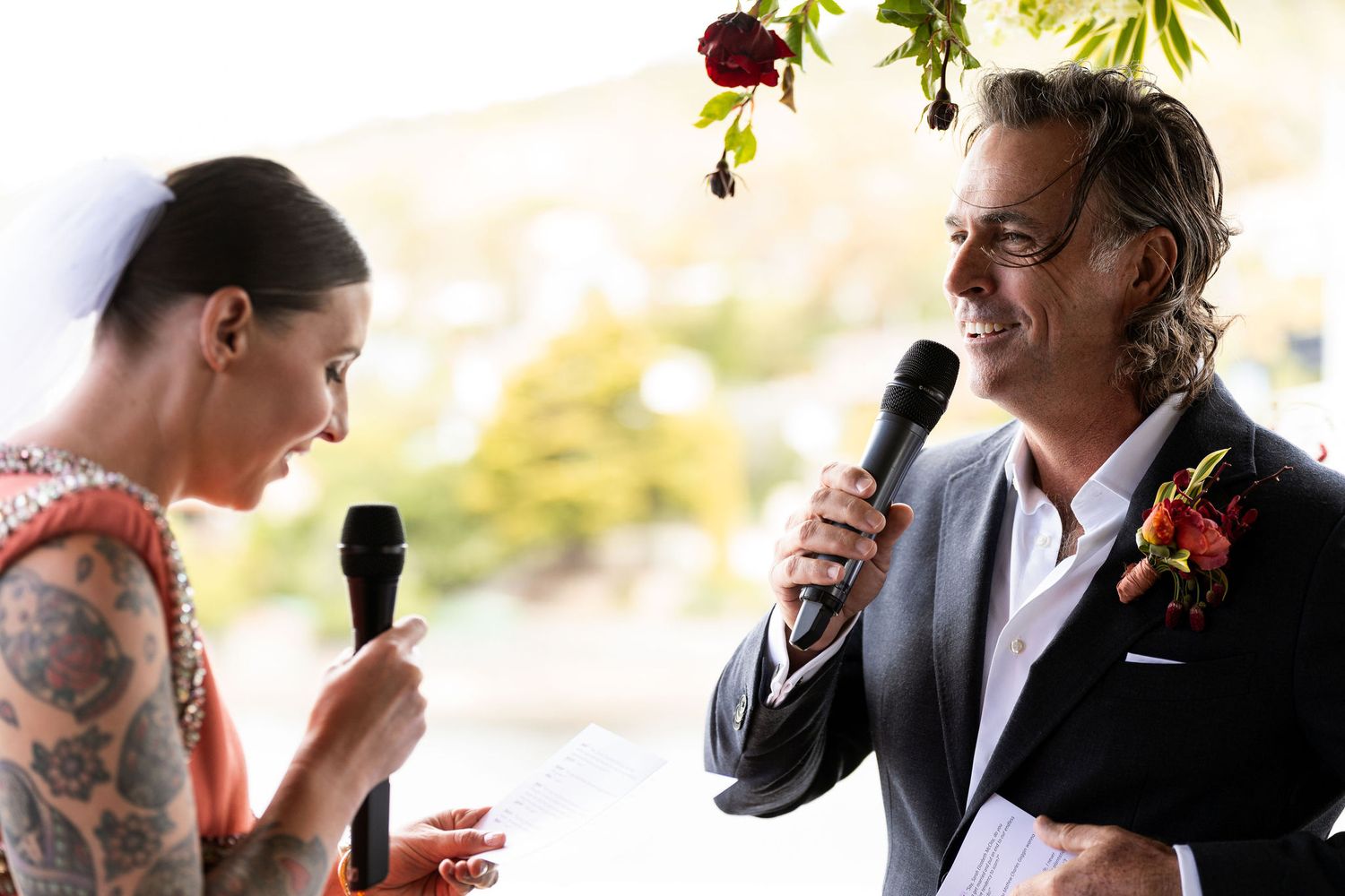 Bride with tattoos and groom in suit exchange wedding vows holding microphones at outdoor ceremony.