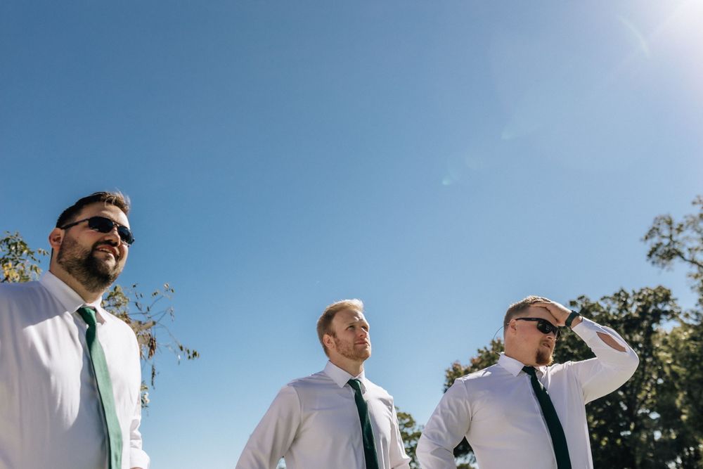 Three groomsmen in matching suits and ties looking off into distance against blue sky at outdoor wedding ceremony in newport news, virginia