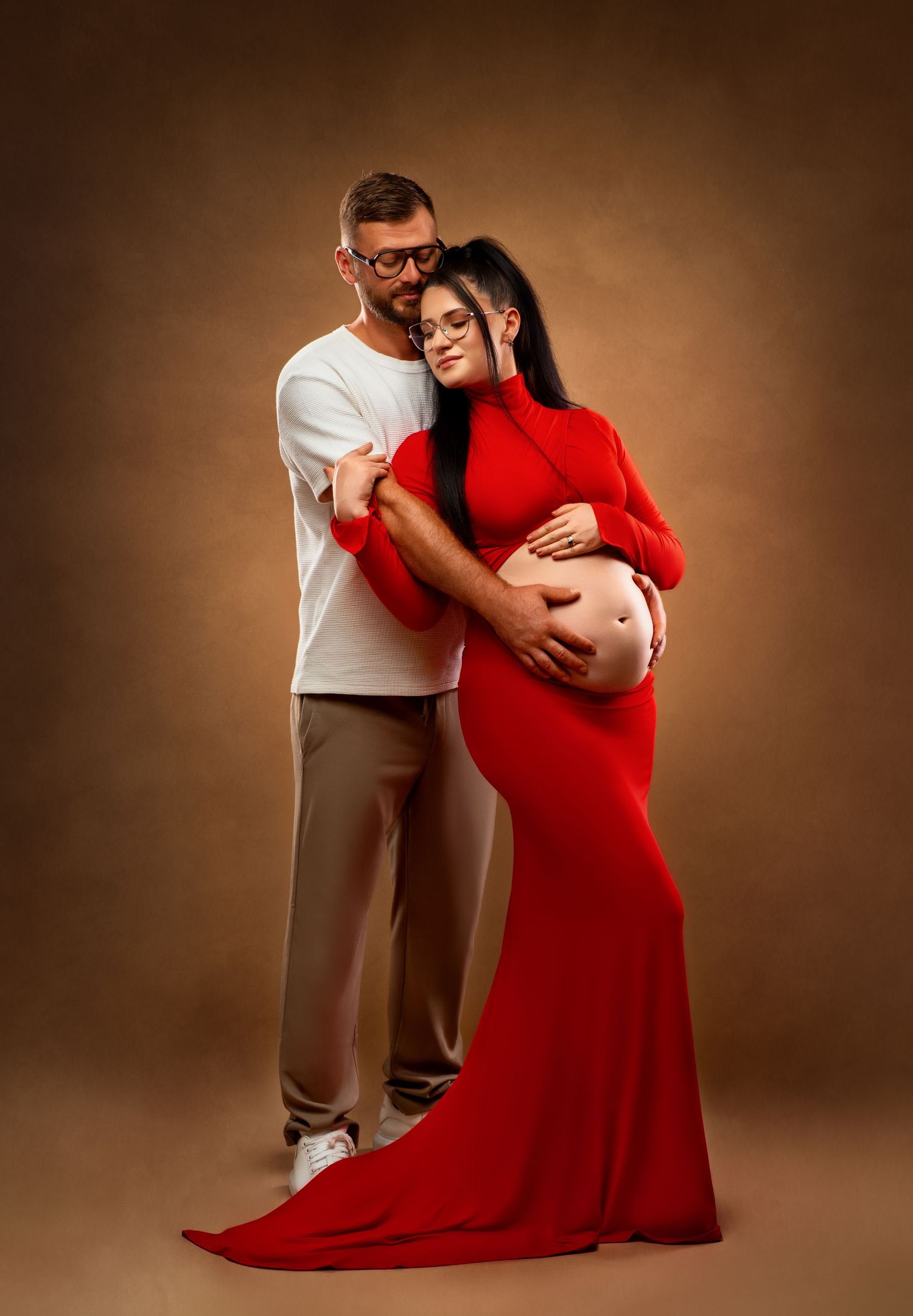 A woman in a flowing red maternity dress embraces her pregnancy bump while standing against a sepia-toned backdrop.