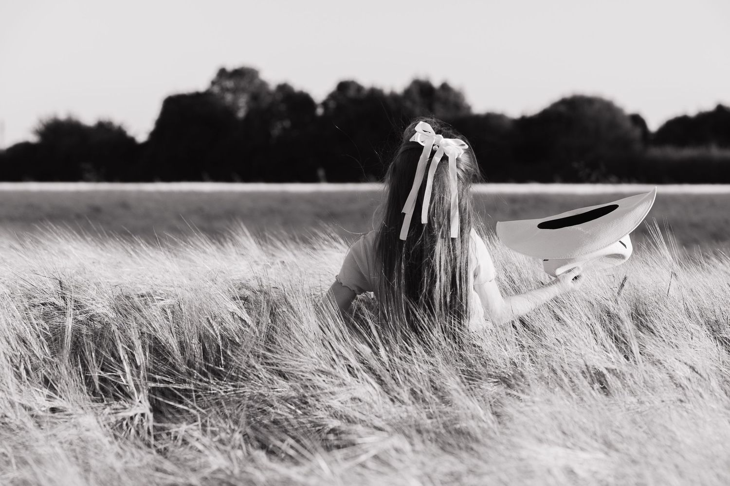 A black and white image of a little girl with her back to the camera, in a wheat field. She is holding a cowboy hat upside down.