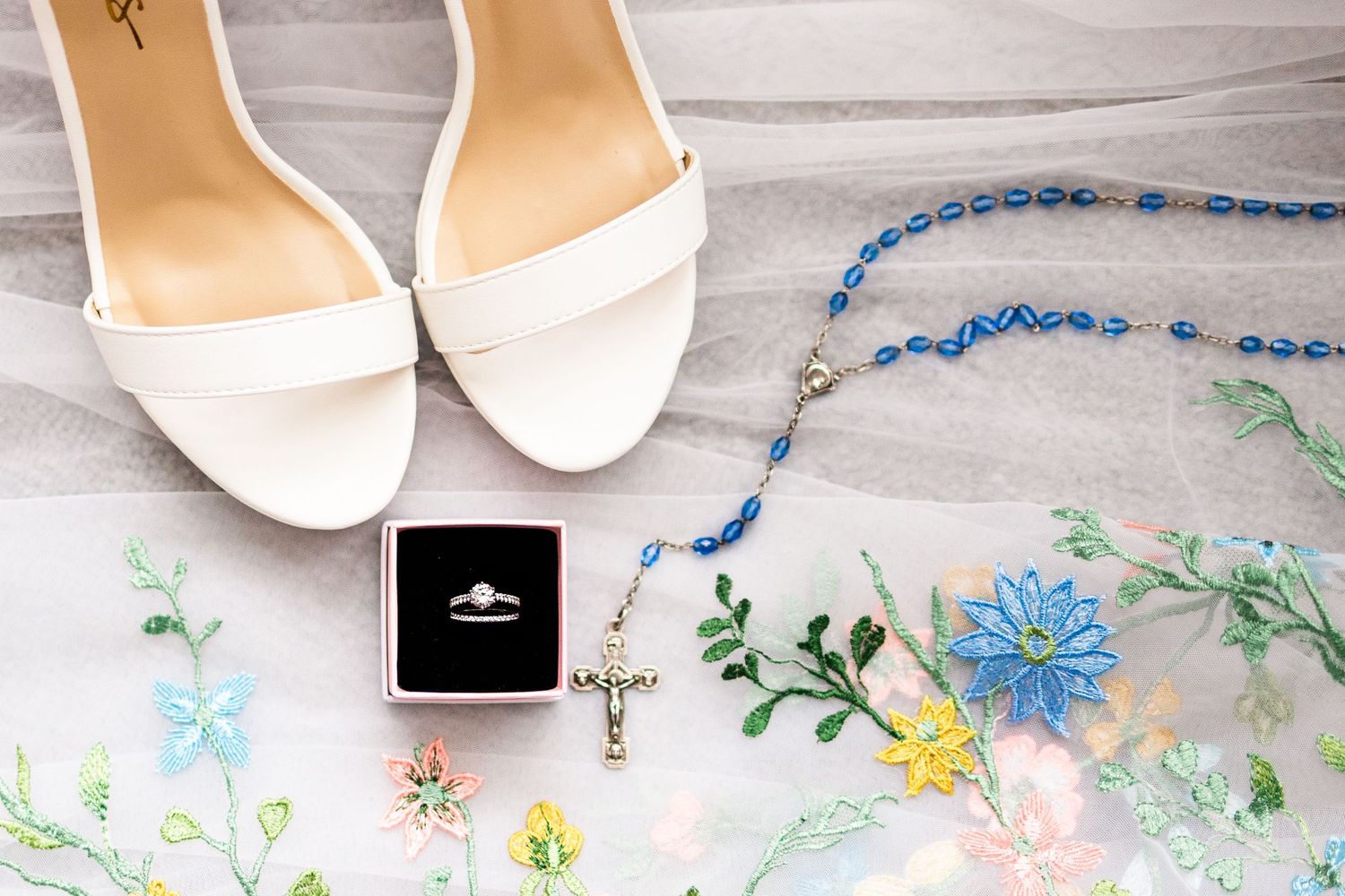 Wedding details including white sandals, a ring box, and blue beaded jewelry arranged with delicate flowers.