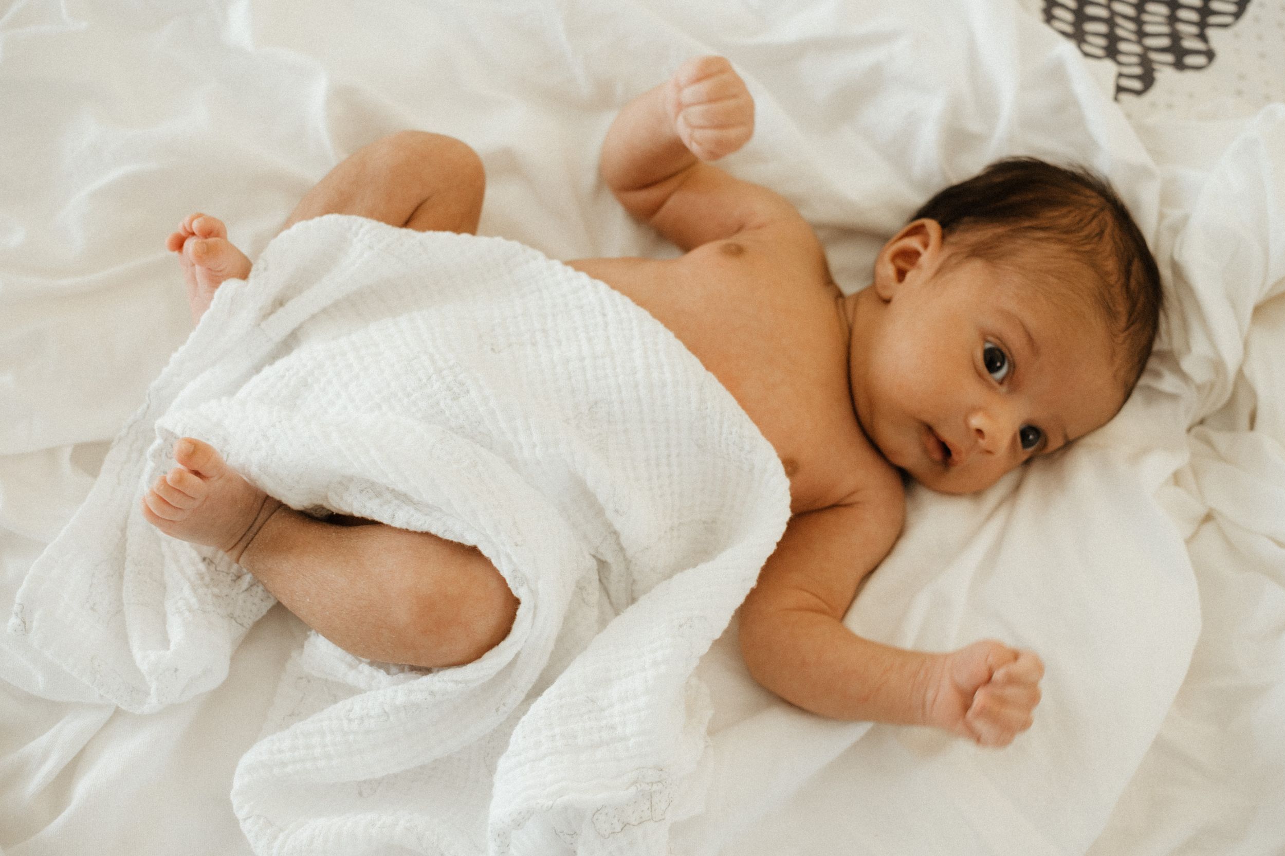 A newborn baby lying on white bedding looking towards the camera.