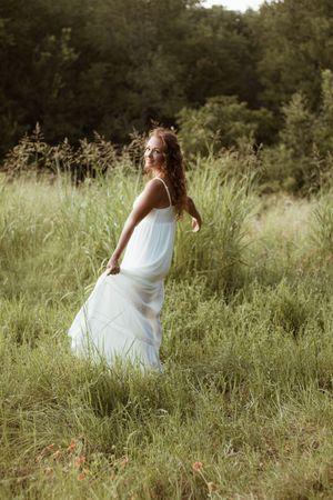 Young woman in flowing white dress standing in lush green field with tall grass and trees in background.