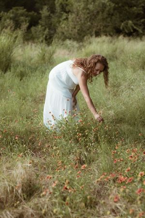Woman in white dress picking wildflowers in a lush green meadow with red blooms on a warm day.
