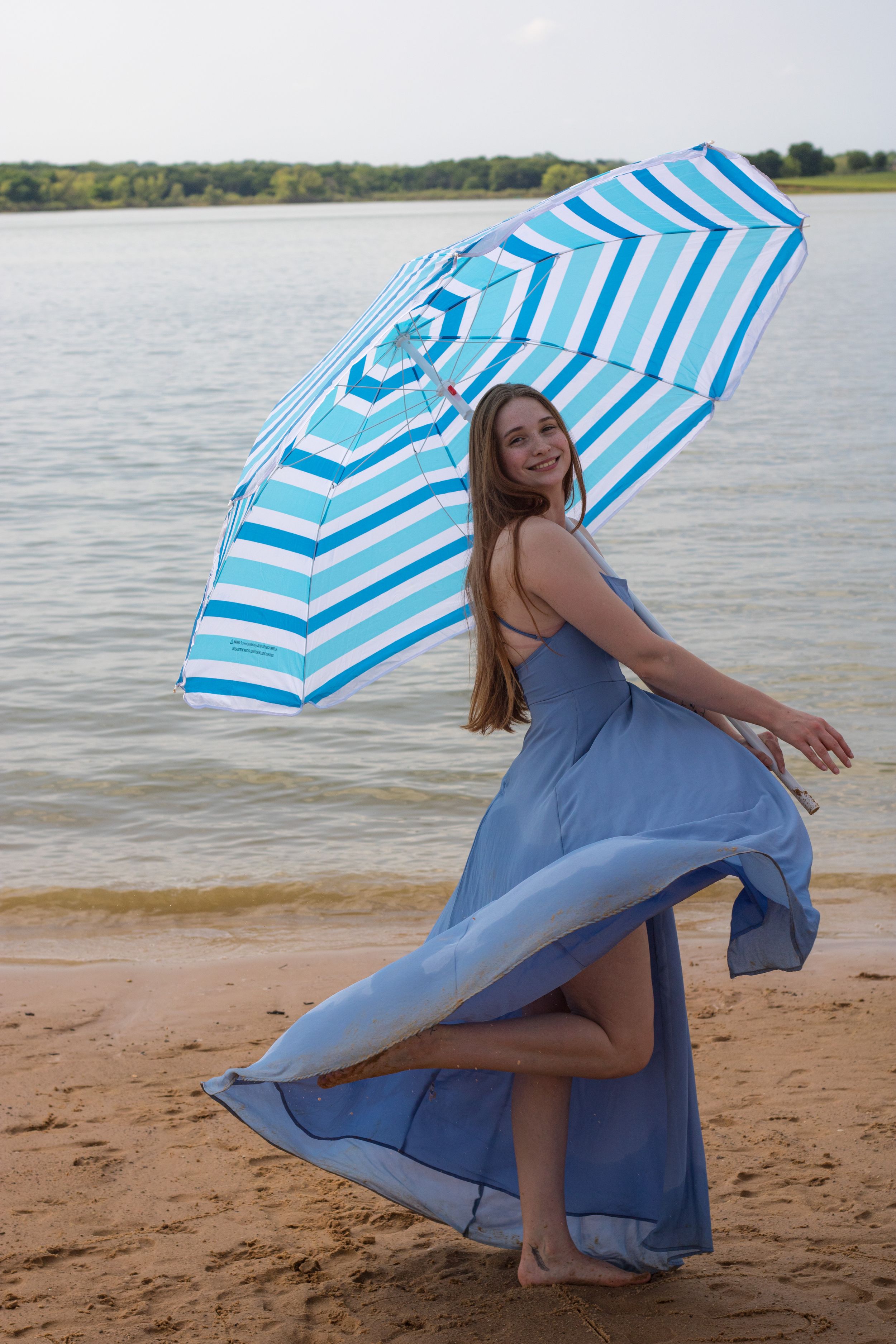 Smiling woman in blue dress holding striped beach umbrella, dancing on sandy shore with lake in background.