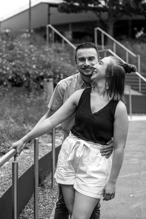 Black and white photo of a young couple smiling and embracing outdoors near stairs and railing.