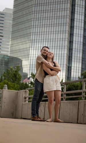A couple embraces on a bridge with modern glass skyscrapers in the background in an urban city setting.
