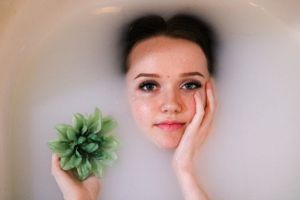 Young woman with freckles relaxing in a milky bath, holding a green succulent flower, resting her cheek on her hand.