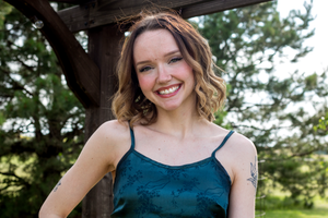 Smiling young woman with curly hair wearing a teal spaghetti strap top outdoors near wooden structure.