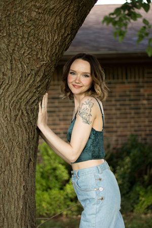 Young woman with tattoo in teal crop top and jeans leaning against tree trunk outdoors near brick house.