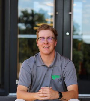 Smiling young man with glasses wearing a gray polo shirt seated outdoors in a professional profile photo.