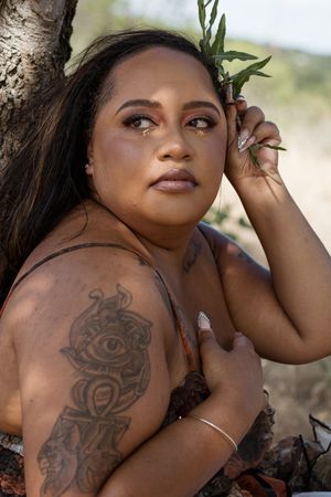Woman with tattoos leaning against a tree holding green leaves, wearing natural makeup in an outdoor setting.