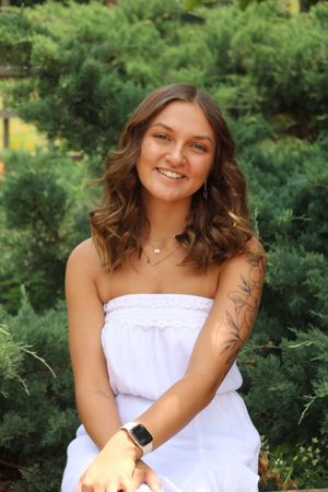 Smiling young woman in white strapless dress with floral arm tattoo, posing outdoors among green evergreen shrubs.