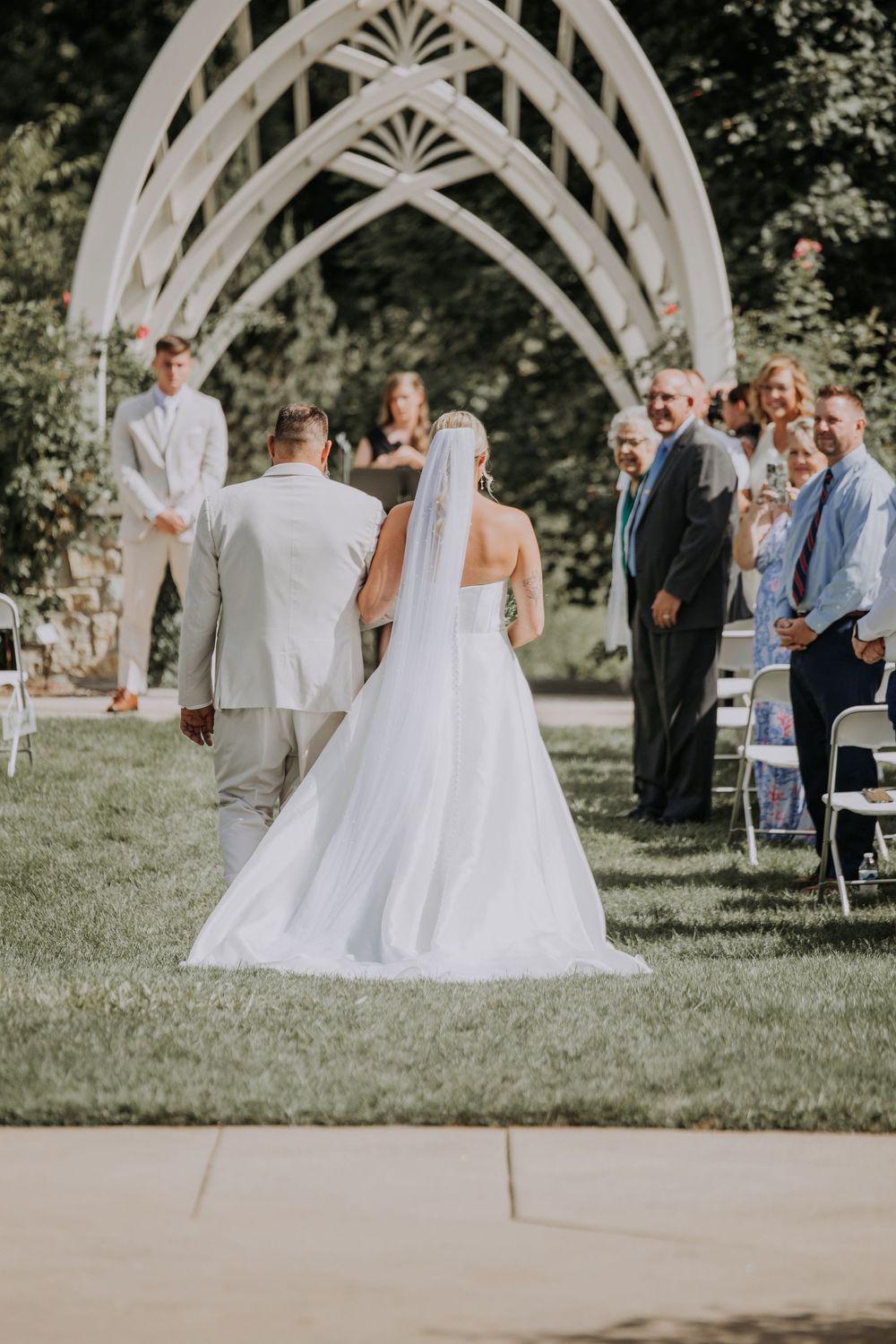 Outdoor wedding ceremony under a white gothic arch with guests seated on the lawn during a sunny summer day.