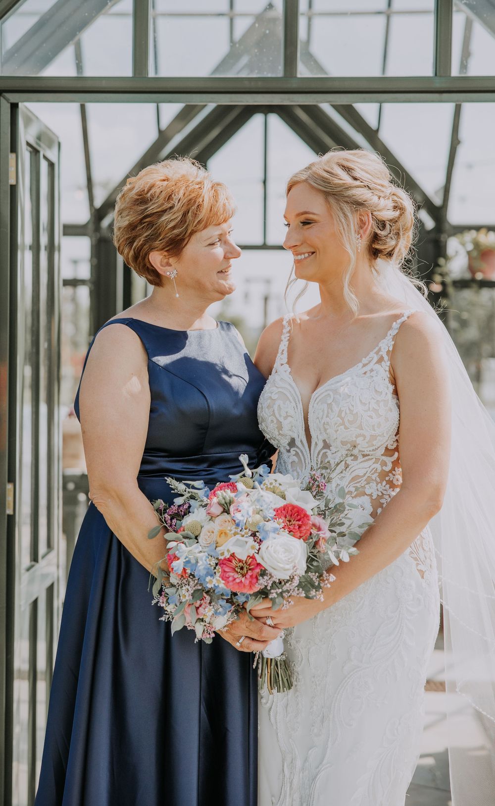 Wedding party members in navy blue dresses share joyful moments with bride in lace gown holding pastel bouquets.