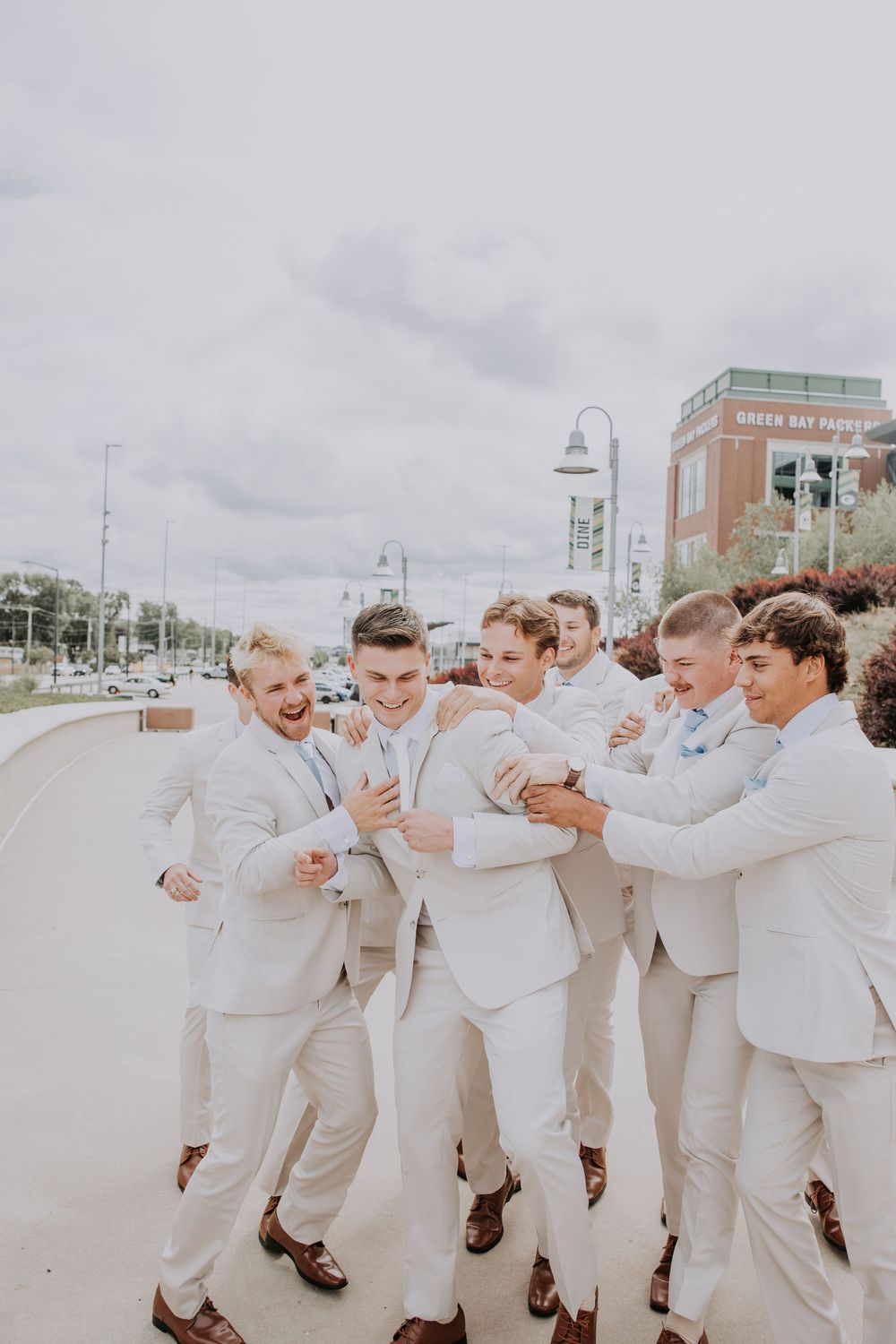 Groomsmen in light beige suits celebrate joyfully together on a cloudy day near a waterfront.