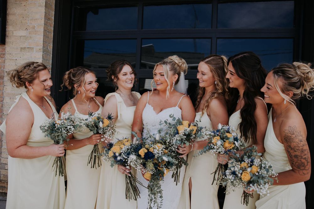 Bridesmaids in light blue dresses stand in line with yellow and white floral bouquets at an outdoor wedding venue.