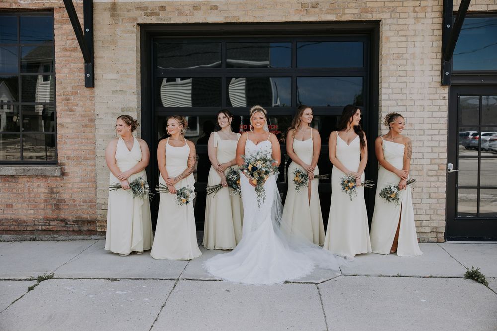Bridesmaids in light yellow dresses stand with bride in white gown against rustic brick wall backdrop.
