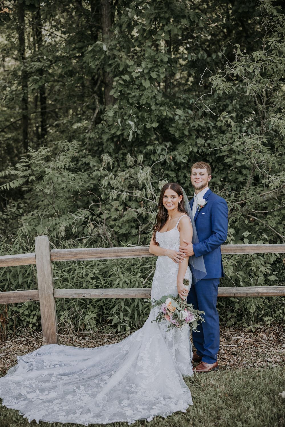 Couple in wedding attire pose together by wooden fence in outdoor garden setting with flowing lace bridal gown.